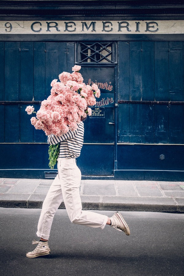 Follow Your Bliss is an image of a girl with a huge bouquet of pink carnations in her arms skipping along the street in front of La Crèmerie in the 6th Arrondissement in Paris.
