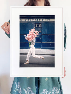Follow Your Bliss is an image of a girl with a huge bouquet of pink carnations in her arms skipping along the street in front of La Crèmerie in the 6th Arrondissement in Paris.